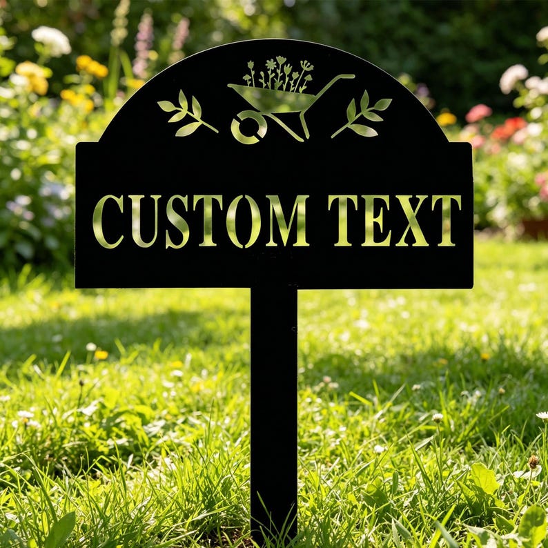 Puede incluir: Letrero de jard&iacute;n de metal negro con las palabras "CUSTOM TEXT" recortadas. La parte superior del letrero presenta una carretilla y un dise&ntilde;o floral. El letrero est&aacute; clavado en un c&eacute;sped con un fondo borroso de flores.
