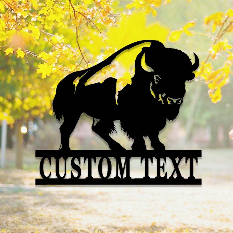 K&ouml;nnte beinhalten: Schwarze Metallsilhouette eines Bisons mit dem Text "CUSTOM TEXT" darunter. Die Silhouette ist vor einem verschwommenen Hintergrund aus Herbstlaub platziert.