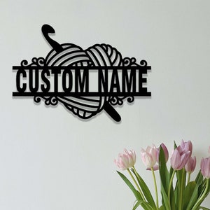 Pode incluir: Uma placa de parede em metal preto com um design de novelo de l&atilde; em forma de cora&ccedil;&atilde;o atravessado por uma agulha de croch&ecirc;. O design inclui uma barra horizontal no centro com o texto CUSTOM NAME em letras mai&uacute;sculas, negrito e sem serifa.