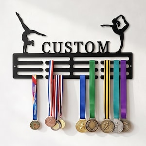 May include: A black metal medal hanger featuring the word CUSTOM in capital letters, flanked by silhouettes of gymnasts in poses. The rack has three horizontal bars with slots for hanging ribbons, currently displaying eight various medals.