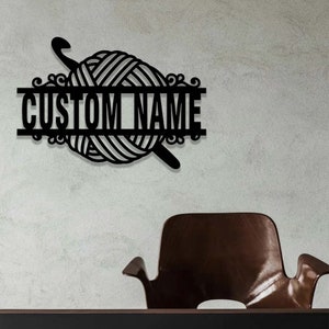 Pode incluir: Uma placa de parede em metal preto com um novelo de l&atilde; central atravessado por uma agulha de croch&ecirc;. Uma barra horizontal atravessa o centro do design, contendo o texto CUSTOM NAME numa fonte negrito, sem serifa e em mai&uacute;sculas.