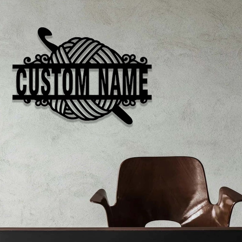 Pode incluir: Uma placa de parede em metal preto com um novelo de l&atilde; central atravessado por uma agulha de croch&ecirc;. Uma barra horizontal atravessa o centro do design, contendo o texto CUSTOM NAME numa fonte negrito, sem serifa e em mai&uacute;sculas.