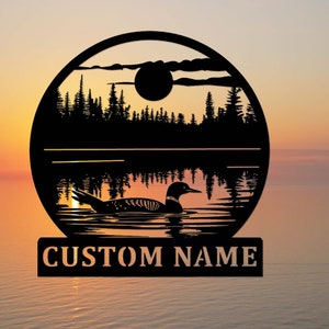 Puede incluir: Arte de pared de metal negro que representa un somorgujo nadando en un lago al atardecer. El diseño incluye una silueta de árboles, un sol y el texto "CUSTOM NAME" en la parte inferior. La obra de arte es circular.