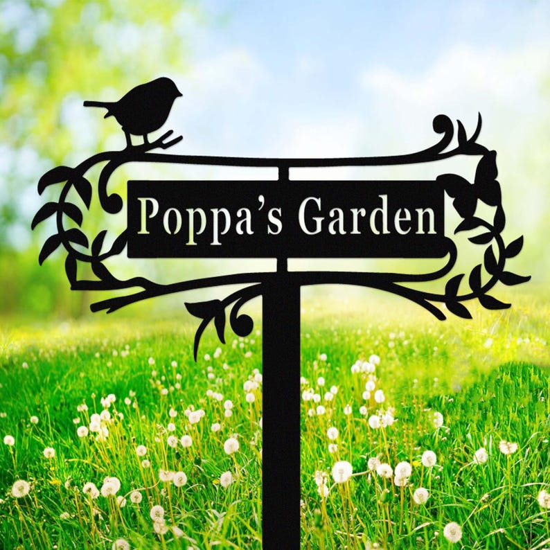 Puede incluir: Letrero de jard&iacute;n de metal negro con el texto "Poppa's Garden". El letrero presenta una silueta de un p&aacute;jaro posado en una rama, con hojas y adornos decorativos. El letrero est&aacute; montado sobre una estaca negra.