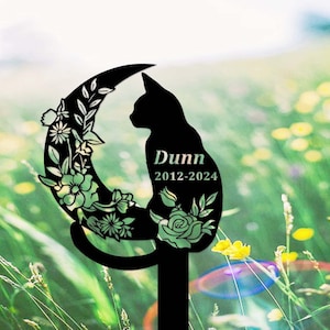 Peut inclure: Silhouette noire en métal d'un chat assis sur un croissant de lune avec des accents floraux. Le nom du chat "Dunn" et les dates "2012-2024" sont gravés sur la silhouette.
