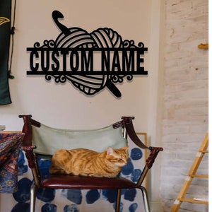 Pode incluir: Uma placa de parede em metal preto com um design de novelo de l&atilde; em forma de cora&ccedil;&atilde;o, uma agulha de croch&ecirc; e uma barra horizontal exibindo o texto CUSTOM NAME em letras mai&uacute;sculas em negrito, com ornamentos decorativos nas bordas.