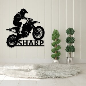 Op de afbeelding: Zwarte metalen silhouet van een motorcrossrijder die springt op een crossmotor met de naam "SHARP" eronder. De silhouet is gemonteerd op een witte muur met een wit, pluche tapijt op de voorgrond.