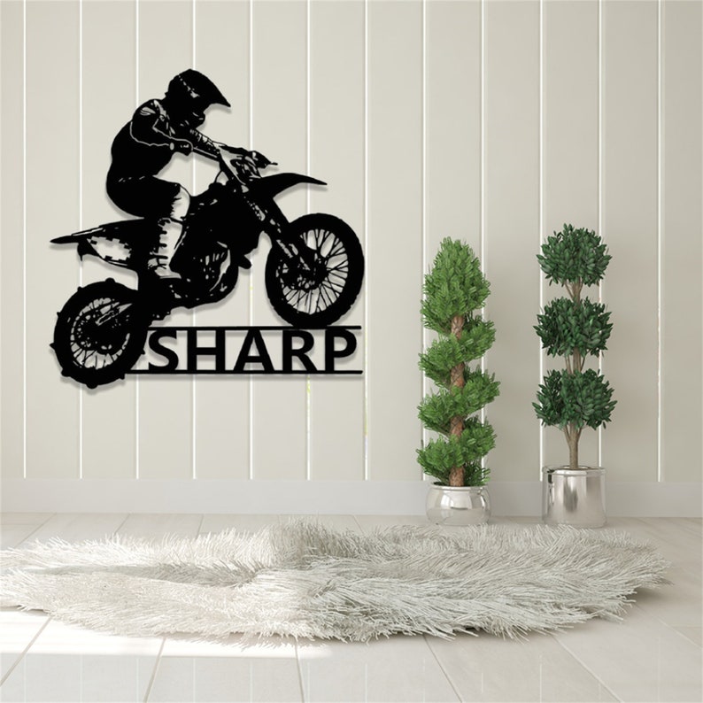 Op de afbeelding: Zwarte metalen silhouet van een motorcrossrijder die springt op een crossmotor met de naam "SHARP" eronder. De silhouet is gemonteerd op een witte muur met een wit, pluche tapijt op de voorgrond.