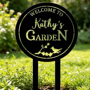 Puede incluir: Letrero de jard&iacute;n de metal negro con el texto "WELCOME TO Kathy's GARDEN" en un dise&ntilde;o circular. Un p&aacute;jaro y una rama floral aparecen en la parte inferior. El letrero est&aacute; colocado en un jard&iacute;n.