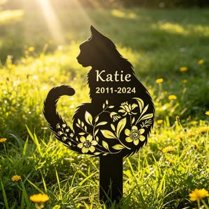 Könnte beinhalten: Schwarzer Metall-Gartenstecker mit Katzensilhouette und floralen Details. Das katzenförmige Denkmal trägt den Namen "Katie" und die Jahreszahlen "2011-2024". Der Stecker befindet sich in einer grasbewachsenen Fläche mit gelben Blumen.