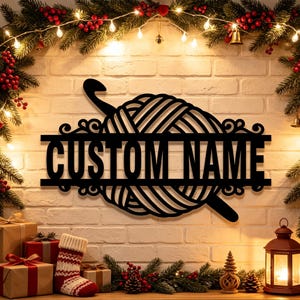 Pode incluir: Uma placa de parede em metal preto com um novelo de l&atilde; e uma agulha de croch&ecirc; inserida. Uma barra horizontal atravessa o centro do novelo, exibindo o texto CUSTOM NAME em letras mai&uacute;sculas em negrito e sem serifa.