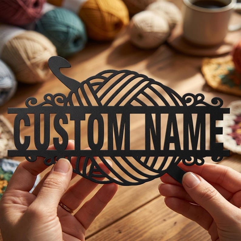 Pode incluir: Uma placa preta cortada a laser com um novelo de l&atilde; estilizado e uma agulha de croch&ecirc; integrada no topo. O design inclui uma barra horizontal no centro com o texto CUSTOM NAME em uma fonte negrito, sem serifa e em letras mai&uacute;sculas.
