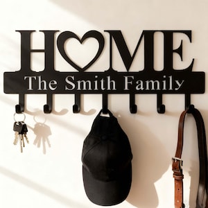 May include: Black metal wall-mounted key and coat rack with the word "HOME" and a heart-shaped design. Below the word "HOME" is the text "The Smith Family". The rack has six hooks, with keys, a hat, and a strap hanging from them.