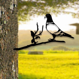Op de afbeelding: Zwarte metalen silhouet van een duif die op een tak met bladeren zit. Het silhouet is bevestigd aan een boomstam.