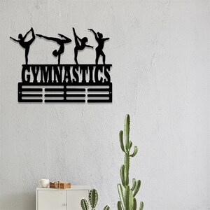 May include: Black metal gymnastics medal display with the word "GYMNASTICS" above a row of medal hooks. The display features four silhouettes of gymnasts in various poses.
