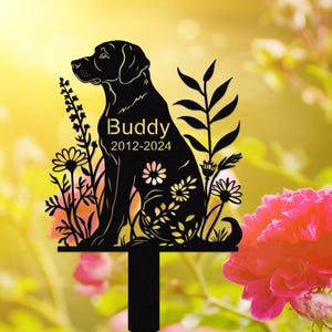 Könnte beinhalten: Schwarze Metallsilhouette eines Hundes, der in einem Blumenfeld sitzt. Der Hund ist ein Labrador Retriever und der Text "Buddy 2012-2024" ist in die Silhouette graviert.