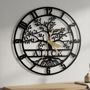 May include: A circular black metal wall clock featuring a cutout tree design with roots and branches. The name ALICE is centered horizontally across the trunk. The clock face has large black numerals from 1 to 12 and gold-colored clock hands.