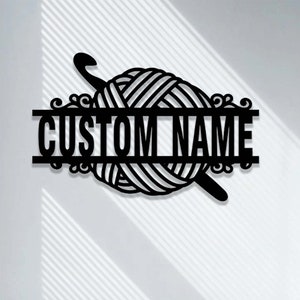 Pode incluir: Uma placa de parede em metal preto com um novelo de l&atilde; central e uma agulha de croch&ecirc; posicionada diagonalmente atr&aacute;s. Uma barra horizontal atravessa o centro do novelo, exibindo o texto CUSTOM NAME em letras mai&uacute;sculas em negrito.