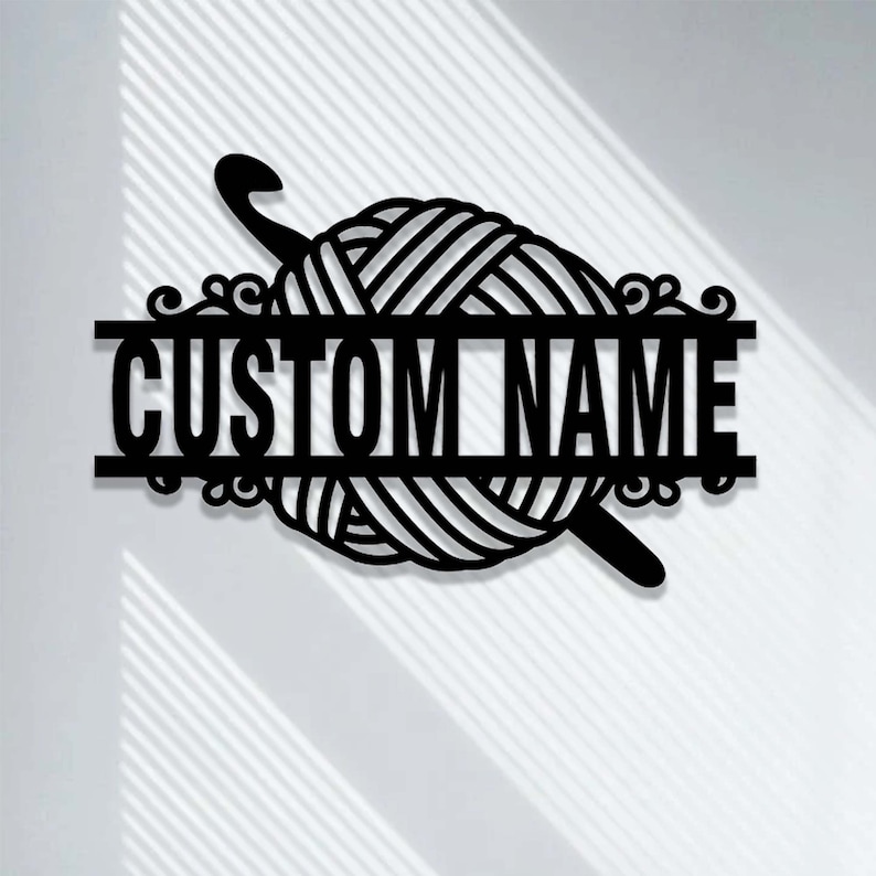 Pode incluir: Uma placa de parede em metal preto com um novelo de l&atilde; central e uma agulha de croch&ecirc; posicionada diagonalmente atr&aacute;s. Uma barra horizontal atravessa o centro do novelo, exibindo o texto CUSTOM NAME em letras mai&uacute;sculas em negrito.