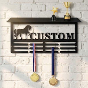 Op de afbeelding: Zwart metalen medaille-display met paardenthema met het woord "CUSTOM" en een paardensilhouet. Inclusief een plank met een sterrenbeker en een gouden beker. Twee medailles hangen eronder.