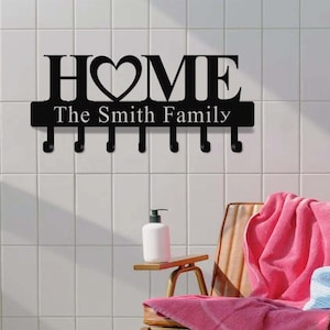 Op de afbeelding: Zwarte metalen wand sleutelhouder met het woord "HOME" en een hartvormig ontwerp in het midden. Onder het woord "HOME" staat de tekst "The Smith Family". De sleutelhouder heeft zes haken.