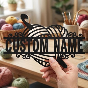 Pode incluir: Uma placa preta cortada a laser com um novelo de l&atilde; estilizado e uma agulha de croch&ecirc;, com o texto CUSTOM NAME centralizado em uma barra horizontal. O design inclui detalhes decorativos em espiral nos lados esquerdo e direito do texto.