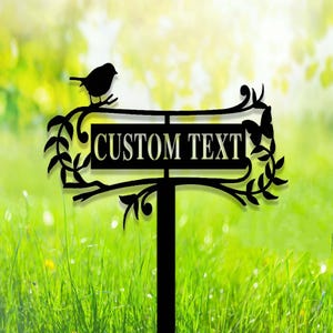 Puede incluir: Se&ntilde;al de jard&iacute;n de metal negro con las palabras "CUSTOM TEXT" en blanco. Una silueta de p&aacute;jaro se posa en la parte superior, con hojas decorativas y una mariposa. La se&ntilde;al est&aacute; en una zona de c&eacute;sped con un fondo verde borroso.