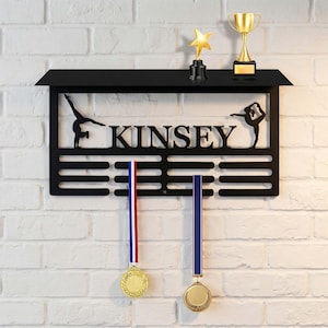 Op de afbeelding: Zwart metalen medaille- en trofeeën display met de naam "KINSEY" uitgesneden. De display heeft silhouetten van gymnasten en bevat twee medailles met rood, wit en blauwe linten, en een gouden ster en trofee.
