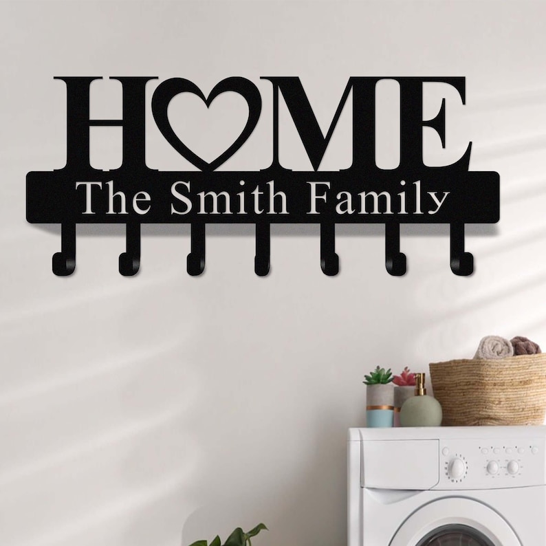 Op de afbeelding: Zwarte metalen wand sleutelhouder met het woord "HOME" en een hartvormig ontwerp. Onder het woord "HOME" staat de tekst "The Smith Family". De sleutelhouder heeft zes haken.