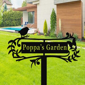 Puede incluir: Letrero de jard&iacute;n de metal negro con el texto "Poppa's Garden" en amarillo. El letrero presenta una silueta de un p&aacute;jaro posado en una rama, con hojas y enredaderas decorativas. El letrero est&aacute; sobre un poste negro, colocado en un c&eacute;sped verde.