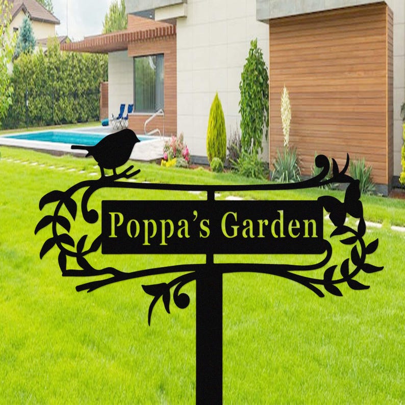 Puede incluir: Letrero de jard&iacute;n de metal negro con el texto "Poppa's Garden" en amarillo. El letrero presenta una silueta de un p&aacute;jaro posado en una rama, con hojas y enredaderas decorativas. El letrero est&aacute; sobre un poste negro, colocado en un c&eacute;sped verde.