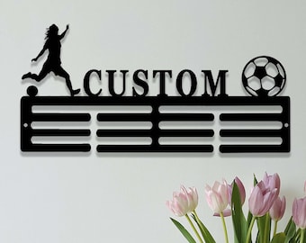 May include: Black metal medal display rack with five rows of hooks. The rack features a silhouette of a soccer player kicking a ball and the word "CUSTOM". A soccer ball is on the right side of the rack.