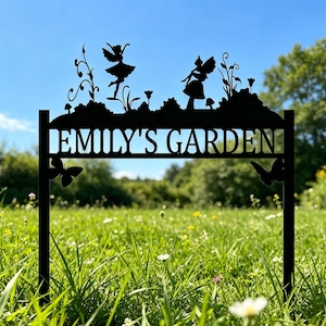 Puede incluir: Letrero de jard&iacute;n de metal negro con el texto "EMILY'S GARDEN". El letrero presenta siluetas de hadas, mariposas y plantas. El letrero est&aacute; colocado en una zona de c&eacute;sped con un cielo azul de fondo.