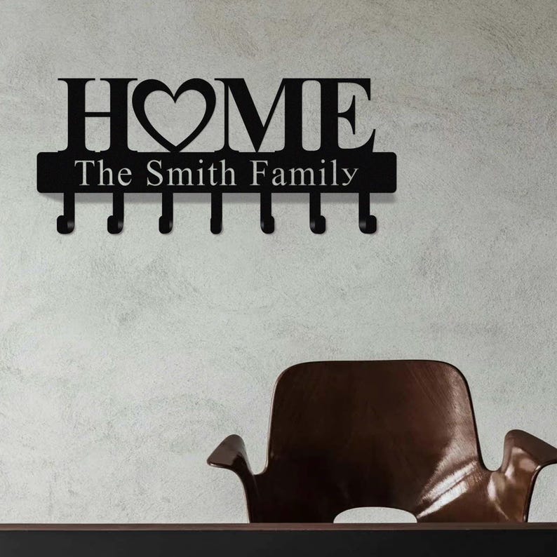 Op de afbeelding: Zwarte metalen wand sleutelhouder met het woord "HOME" en een hartvormig symbool. Onder het woord "HOME" staat de tekst "The Smith Family". De sleutelhouder heeft zes haken om sleutels of andere kleine items op te hangen.