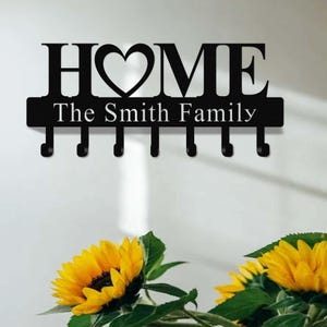 Op de afbeelding: Zwarte metalen sleutelhouder met het woord "HOME" en een hartvormig ontwerp. Onder het woord "HOME" staat de tekst "The Smith Family". De sleutelhouder heeft zes haken. Gele zonnebloemen staan op de voorgrond.