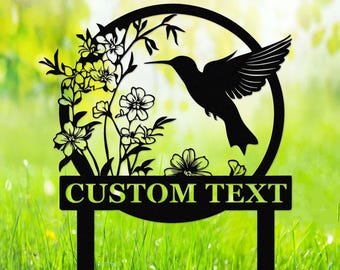 Puede incluir: Estaca de jardín de metal negro en forma de colibrí con un diseño circular que presenta un colibrí en vuelo y acentos florales. La estaca tiene un letrero rectangular que dice "CUSTOM TEXT".