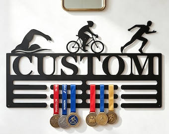 May include: A black metal wall-mounted medal hanger featuring silhouettes of a swimmer, a cyclist, and a runner above the word CUSTOM in bold capital letters. The display includes six horizontal bars for hanging medals with ribbons attached.