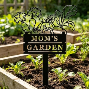 Puede incluir: Letrero de jard&iacute;n de metal negro con las palabras "MOM'S GARDEN" en una fuente decorativa. El letrero presenta siluetas recortadas de verduras, incluyendo zanahorias, una calabaza y verduras de hoja verde. El letrero est&aacute; colocado en un parterre con plantas en crecimiento.