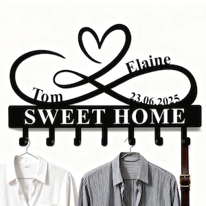 Op de afbeelding: Zwarte metalen wandkapstok met de tekst "SWEET HOME" en zes haken. Het ontwerp bevat een oneindigheidssymbool met een hart, de namen "Tom" en "Elaine" en de datum "23.06.2025". Twee shirts en een riem hangen aan de haken.