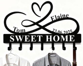 Op de afbeelding: Zwarte metalen wandkapstok met de tekst "SWEET HOME" en zes haken. Het ontwerp bevat een oneindigheidssymbool met een hart, de namen "Tom" en "Elaine" en de datum "23.06.2025". Twee shirts en een riem hangen aan de haken.