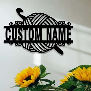 Pode incluir: Uma placa de parede em metal preto com um novelo de l&atilde; e uma agulha de croch&ecirc;. O design inclui uma barra horizontal no centro com o texto CUSTOM NAME em letras mai&uacute;sculas em negrito, acentuado por ornamentos decorativos nas laterais.