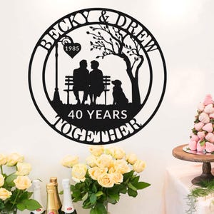 Op de afbeelding: Zwart metalen wandkunst met een stel en een hond op een bank onder een boom. De namen "Becky & Drew" en het jaar "1985" worden weergegeven, met de tekst "40 YEARS TOGETHER" in een cirkelvormig ontwerp. Romantisch cadeau.