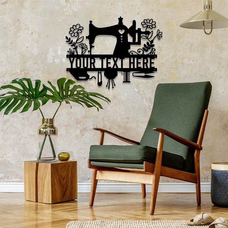 May include: Black metal sewing machine wall art with floral and leaf accents. The text "YOUR TEXT HERE" is displayed below the sewing machine. The wall art is displayed on a textured wall, with a green chair and wooden side table in the foreground.