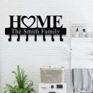 Op de afbeelding: Zwarte metalen wand sleutelhouder met het woord "HOME" en een hartvormige "O". Daaronder staat "The Smith Family". Het heeft zes haken voor sleutels. De achtergrond is een witte bakstenen muur.