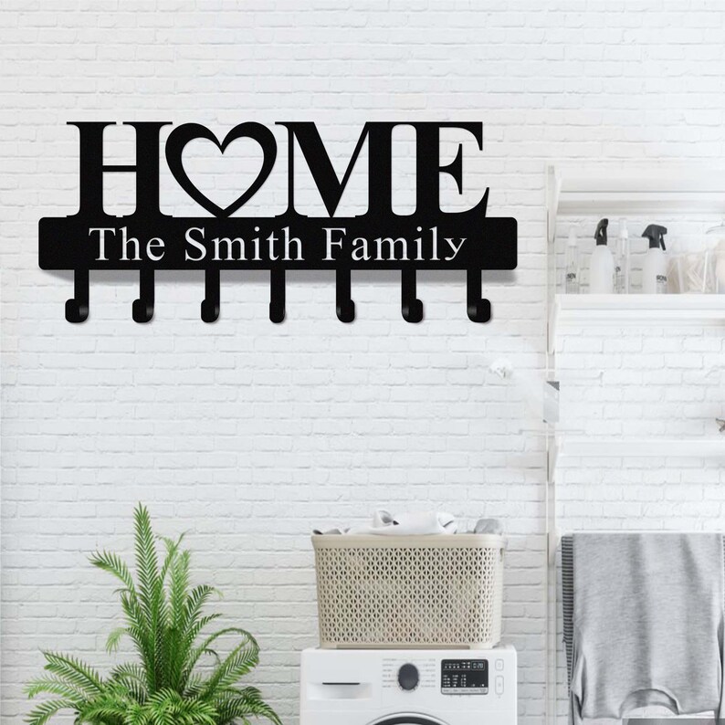 Op de afbeelding: Zwarte metalen wand sleutelhouder met het woord "HOME" en een hartvormige "O". Daaronder staat "The Smith Family". Het heeft zes haken voor sleutels. De achtergrond is een witte bakstenen muur.