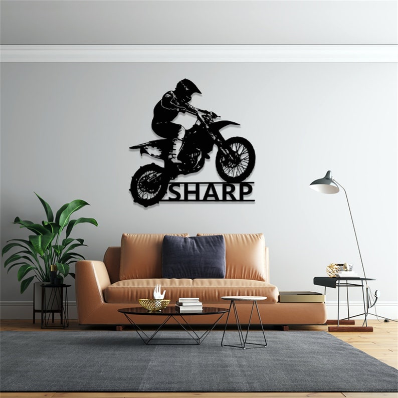 Op de afbeelding: Zwarte metalen silhouet van een motorcrossrijder die springt op een crossmotor met de naam "SHARP" eronder. De silhouet is gemonteerd op een lichtgrijze muur boven een bruine leren bank.