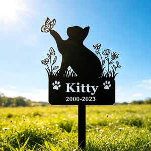 May include: Black metal pet memorial stake with a cat silhouette and butterfly detail. The sign reads "Kitty" and the years 2000-2023, with paw prints. The stake is placed in a grassy field against a blue sky, suitable for a pet's grave.