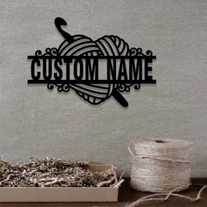 Pode incluir: Uma placa de parede em metal preto com um novelo de l&atilde; em forma de cora&ccedil;&atilde;o e uma agulha de croch&ecirc;. O design inclui uma barra horizontal no centro com o texto CUSTOM NAME em letras mai&uacute;sculas em negrito, rodeado por detalhes decorativos.