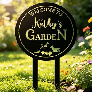 Puede incluir: Letrero de jard&iacute;n de metal negro con el texto "WELCOME TO Kathy's GARDEN" en un dise&ntilde;o circular. El letrero presenta una silueta de p&aacute;jaro y rama floral. El letrero est&aacute; colocado en un jard&iacute;n con hierba verde y flores coloridas.