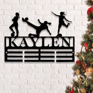 以下が含まれることがあります： 「KAYLEN」という名前が書かれた黒い金属製のメダルディスプレイ。メダルを収納できるスロットが12個あり、ディスプレイの上にはサッカーをしている3人の女性アスリートのシルエットが描かれている。